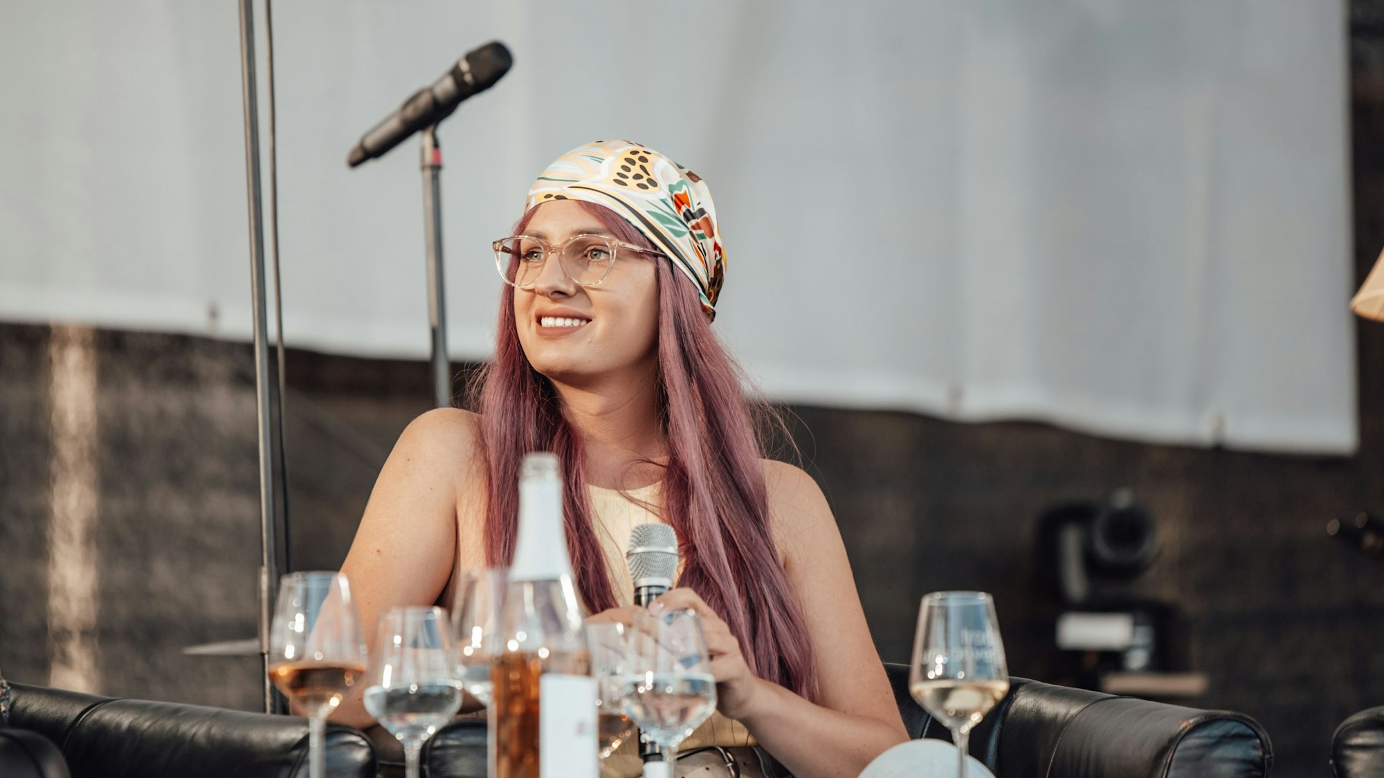 Leni Bolt mit langen pinken Haaren und großer Brille an einem Tisch, sie hält ein Mikro in der Hand.