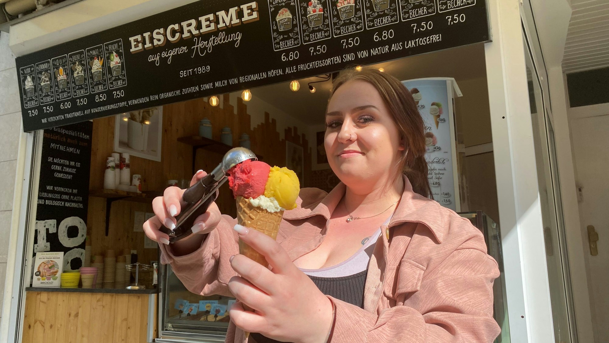 Eine Frau mit einem Nasenring packt vor einer Eisdiele eine Kugel mit einer Eiskelle auf eine Waffel.
