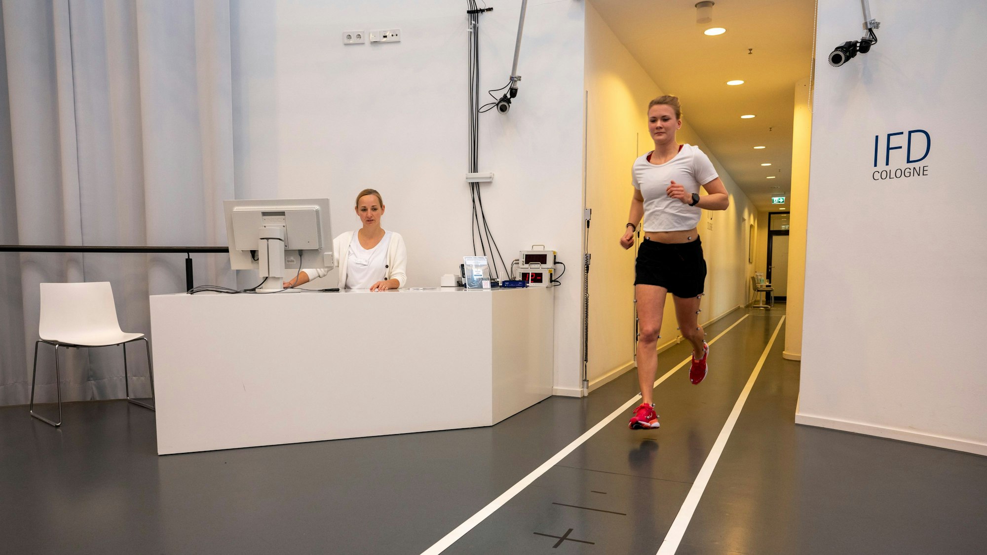 Eine mit Sensoren ausgestattete junge Frau läuft auf einer Labor-Bahn. Eine Labor-Mitarbeiterin sitzt an einem Computer und begleitet den Versuch.