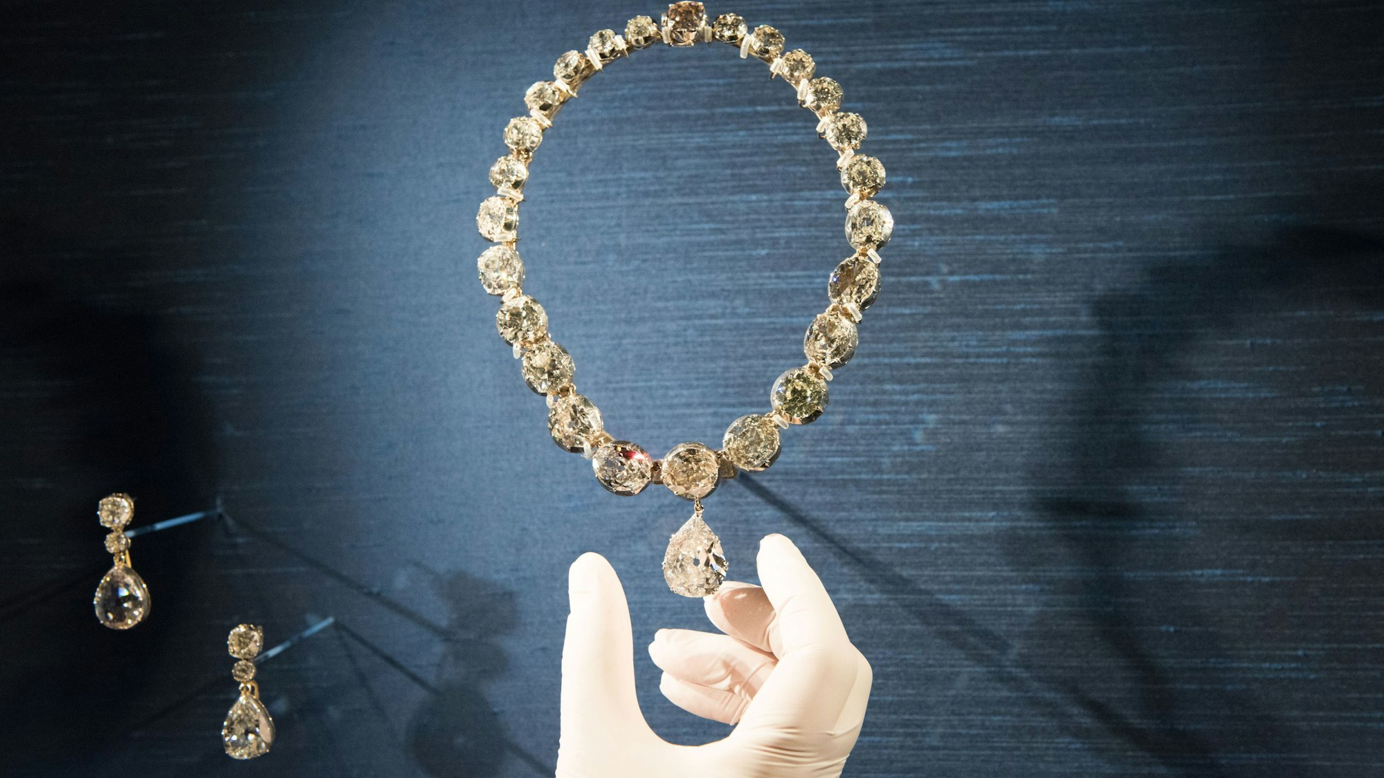 Krönungs-Halskette und -Ohrringe bei einer Ausstellung im Jahre 2022. Eine Hand mit Handschuhen greift nach der Kette.