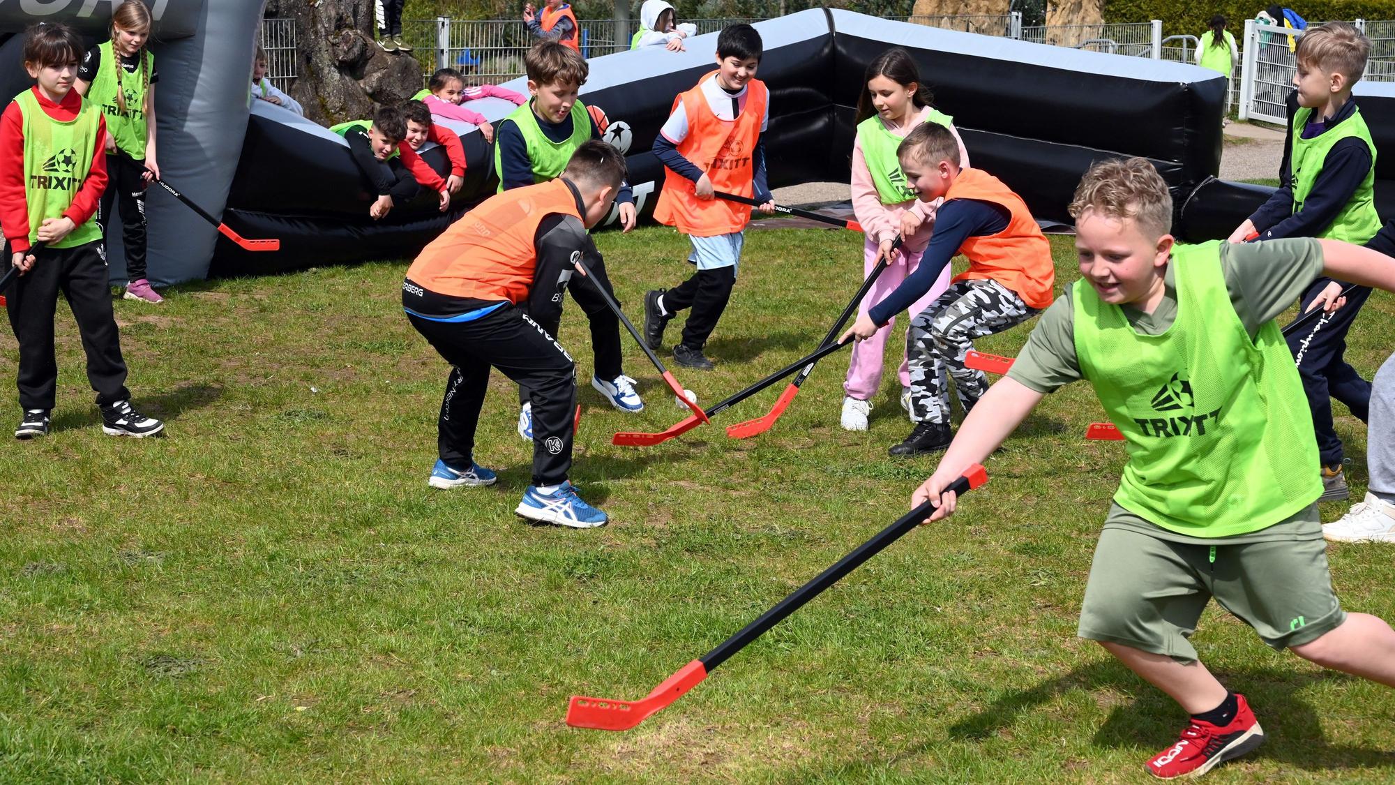 Kinder in orangefarbenen und gelben Leibchen spielen auf einem Feld auf einer Wiese Hockey.