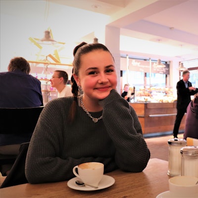 Influencerin Florentine alias „Flobroo“ aus Köln sitzt in einem Lokal, sie trägt einen geflochtenen, dunkelbraunen Pferdeschwanz und eine silberne Gliederkette.