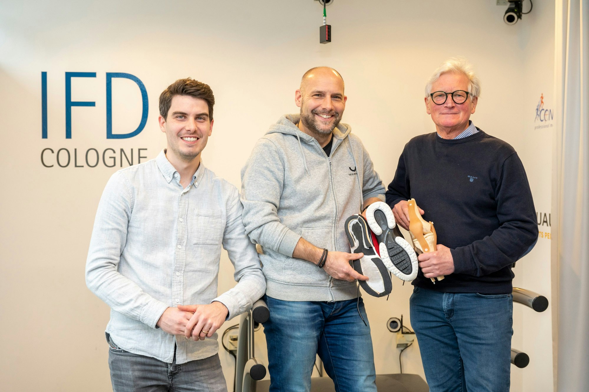 Andre Kriwet, Christian Arens und Gerd-Peter Brüggemann sind im Gruppenporträt vor dem Logo des Test-Labors zu sehen. Arens hält zwei neue Laufschuh-Modelle in der Hand und zeigt die Sohlen.