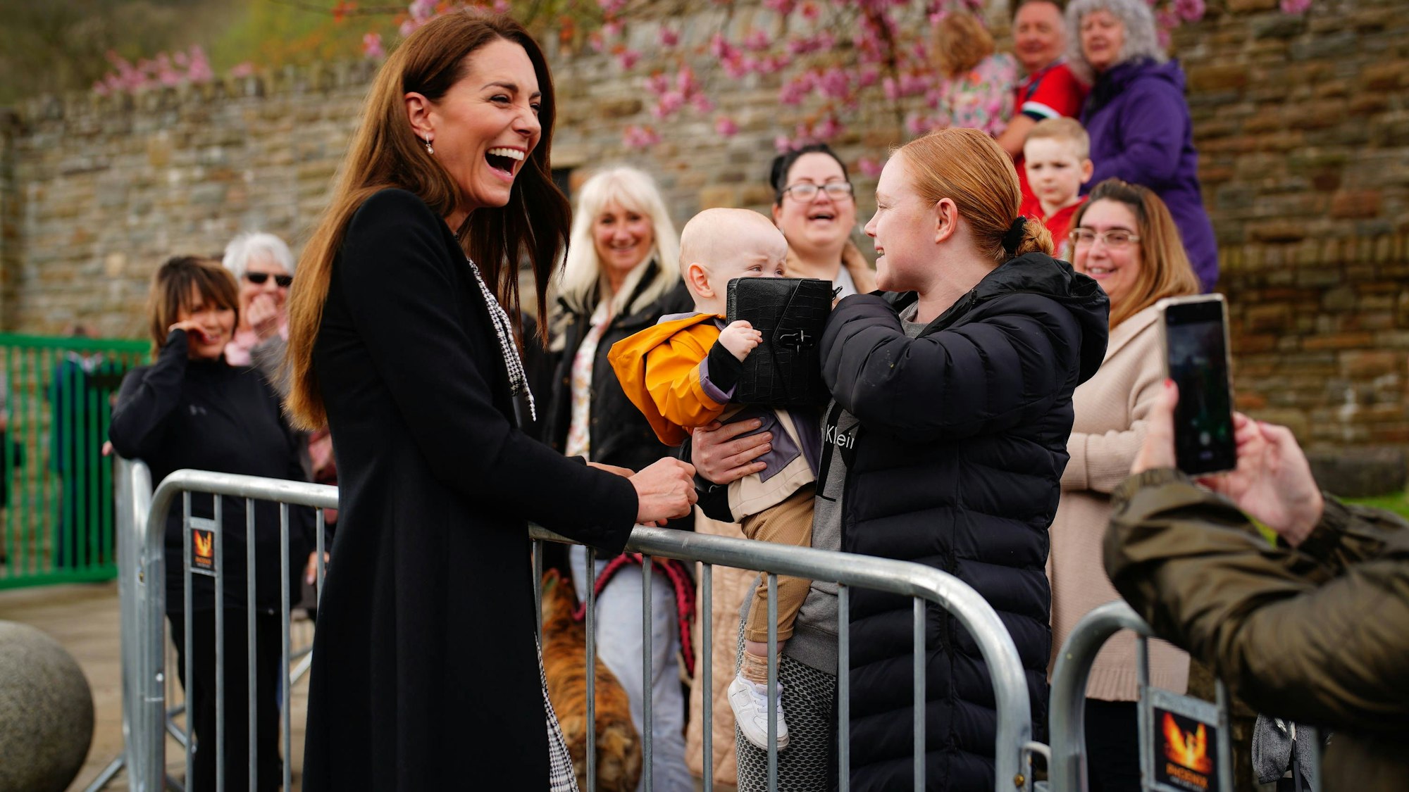Kate Middleton lacht, während ein Baby mit ihrer Handtasche spielt. Seine Mutter hält das Baby auf dem Arm.