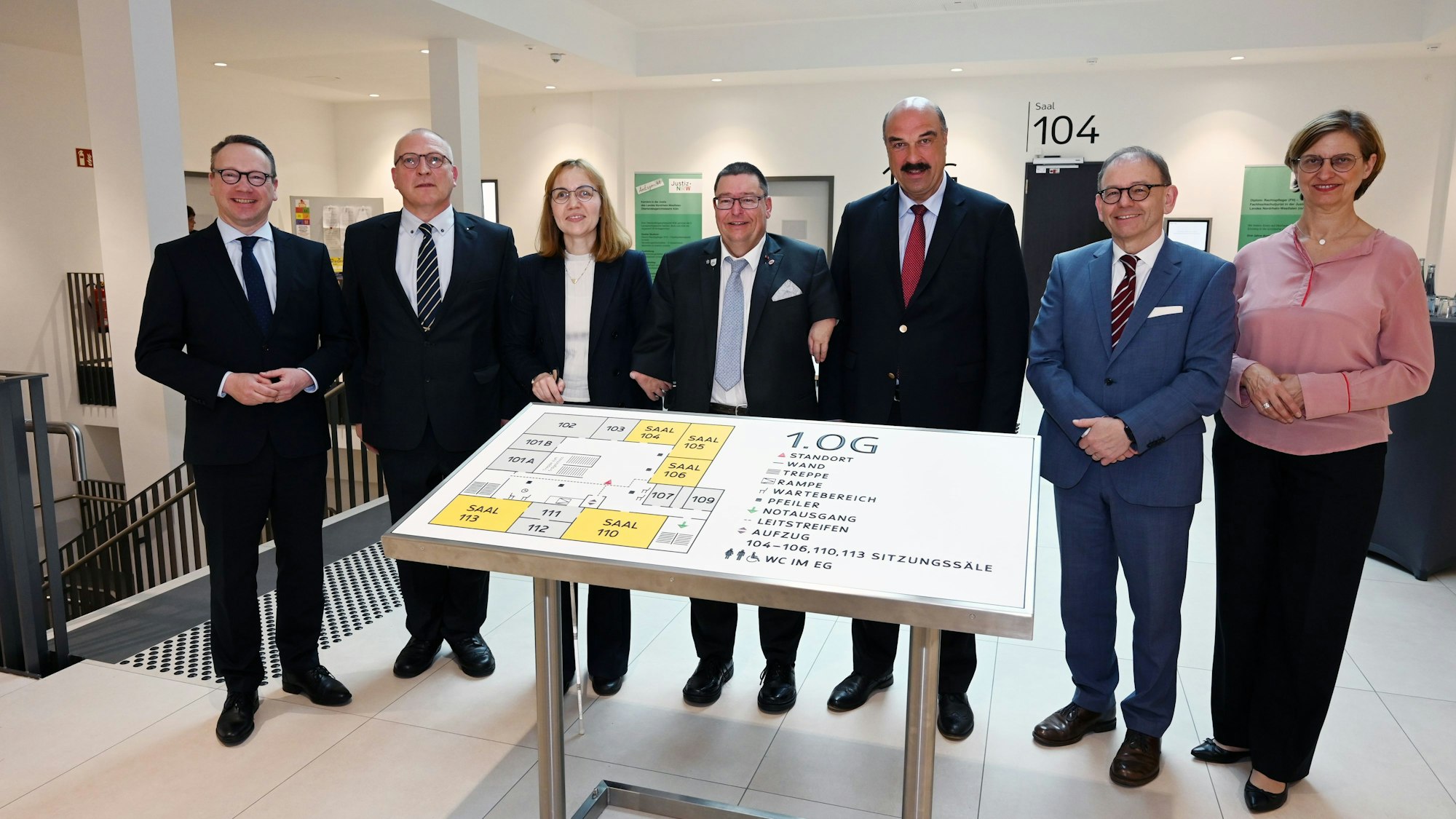 Mehrere Männer und Frauen stehen in einem Gerichtsgebäude vor einer großen Tafel für Sehbehinderte.