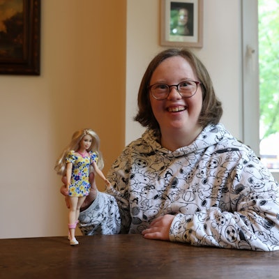 Die Kölnerin Natalie Dedreux lacht in die Kamera und hält die neue Barbie mit Down-Syndrom in ihrer rechten Hand.