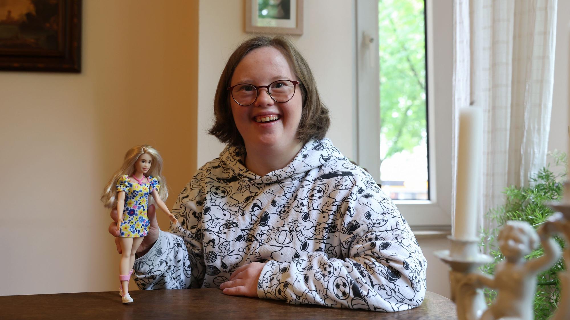 Die Kölnerin Natalie Dedreux lacht in die Kamera und hält die neue Barbie mit Down-Syndrom in ihrer rechten Hand.