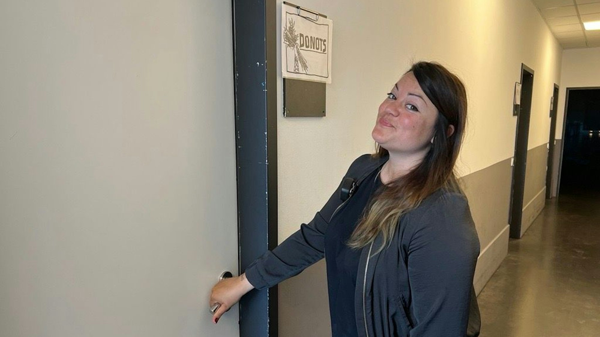 Tourmanagerin Maya Sontheimer vor dem Backstage-Raum der Donots im Palladium in Köln. Neben der Tür hängt ein Schild mit der Aufschrift Donots. Sontheimer drückt die Klinke runter und lächelt in die Kamera.