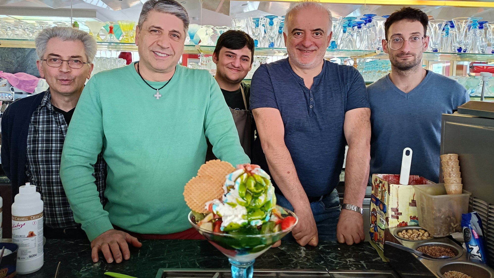 Das Team vom Eiscafé Marino (von links Domenico Marino, Francesco Marino, Sepehr Alidostie, Gennaro Madeo und Tanarico Andrea) blickt fröhlich in die Kamera. Im Vordergrund des Bildes ist ein großer Eisbecher namens „Coppa Fantasia“ zu sehen.
