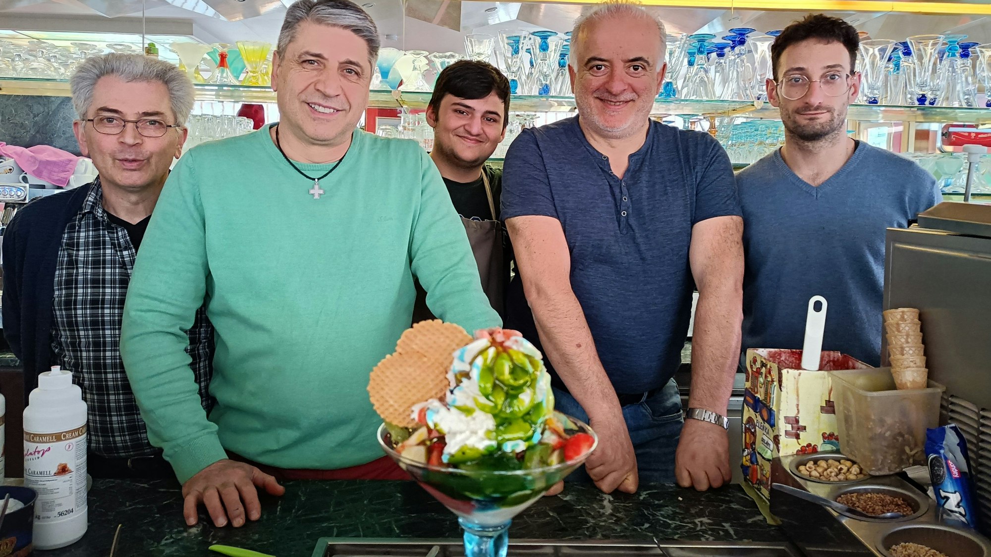 Das Team vom Eiscafé Marino (von links Domenico Marino, Francesco Marino, Sepehr Alidostie, Gennaro Madeo und Tanarico Andrea) blickt fröhlich in die Kamera. Im Vordergrund des Bildes ist ein großer Eisbecher namens „Coppa Fantasia“ zu sehen.