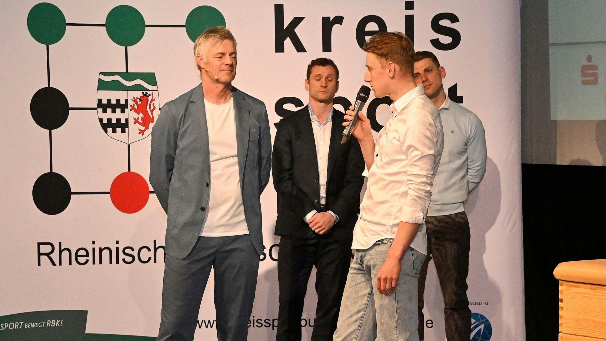 Ein junger Mann mit Mikro sprocht vor einer Gruppe von Menschen vor einer Aufstellwand des Kreissportbundes Rheinisch-Bergischer Kreis.