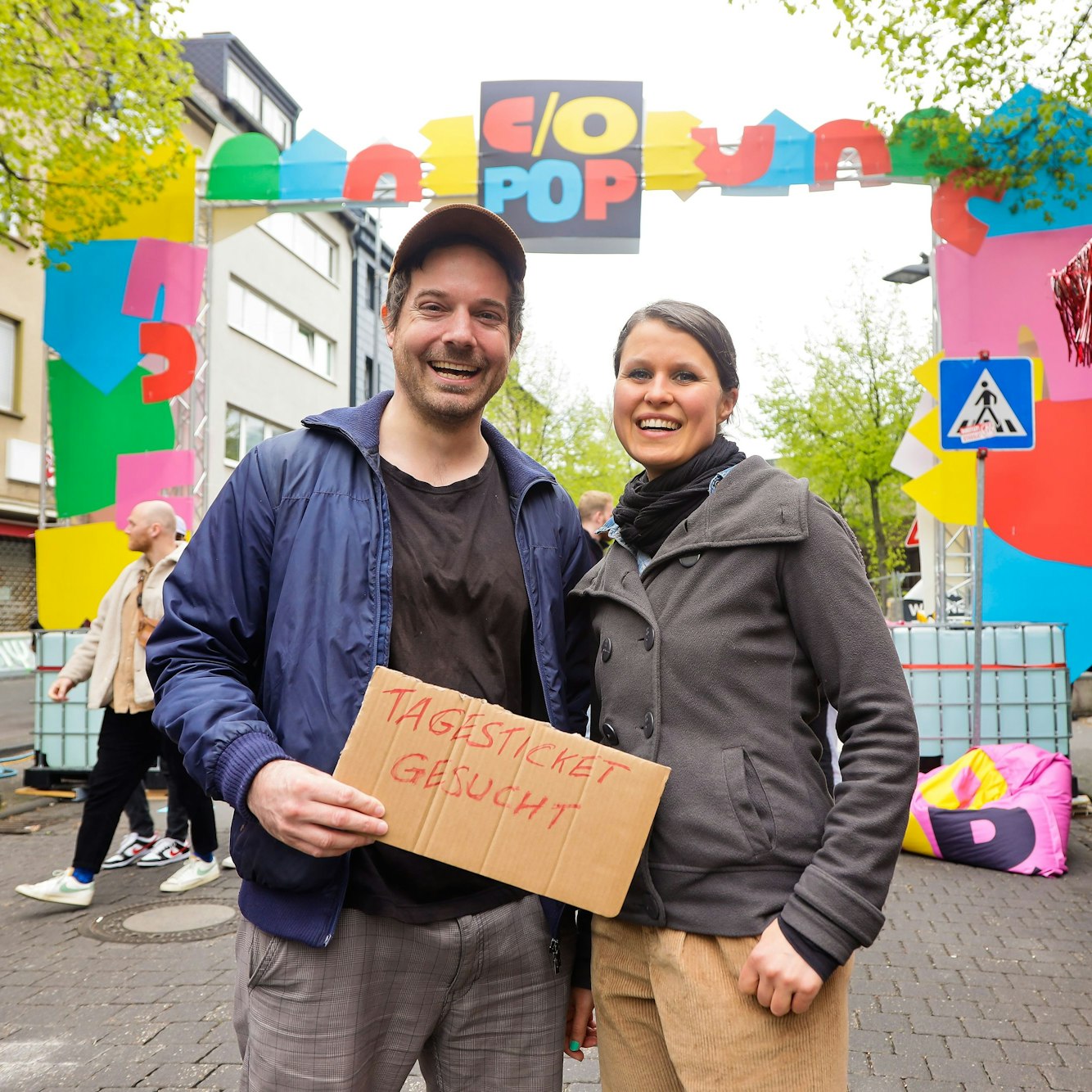 Ein Mann und eine Frau vor der Torbogen der C/o-Pop, sie halten ein Pappschild hoch, auf dem "Tagesticket gesucht" steht.