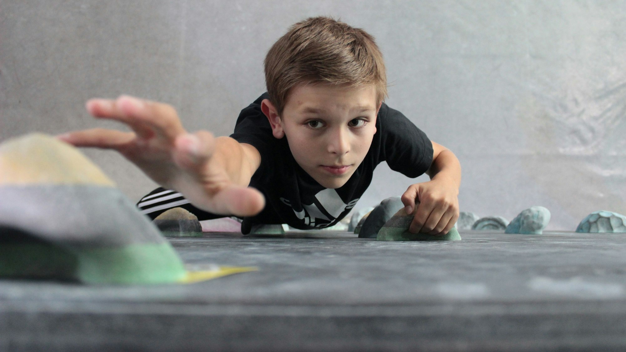 Ein Junge klettert an einer Boulderwand nach oben auf die Kamera zu.