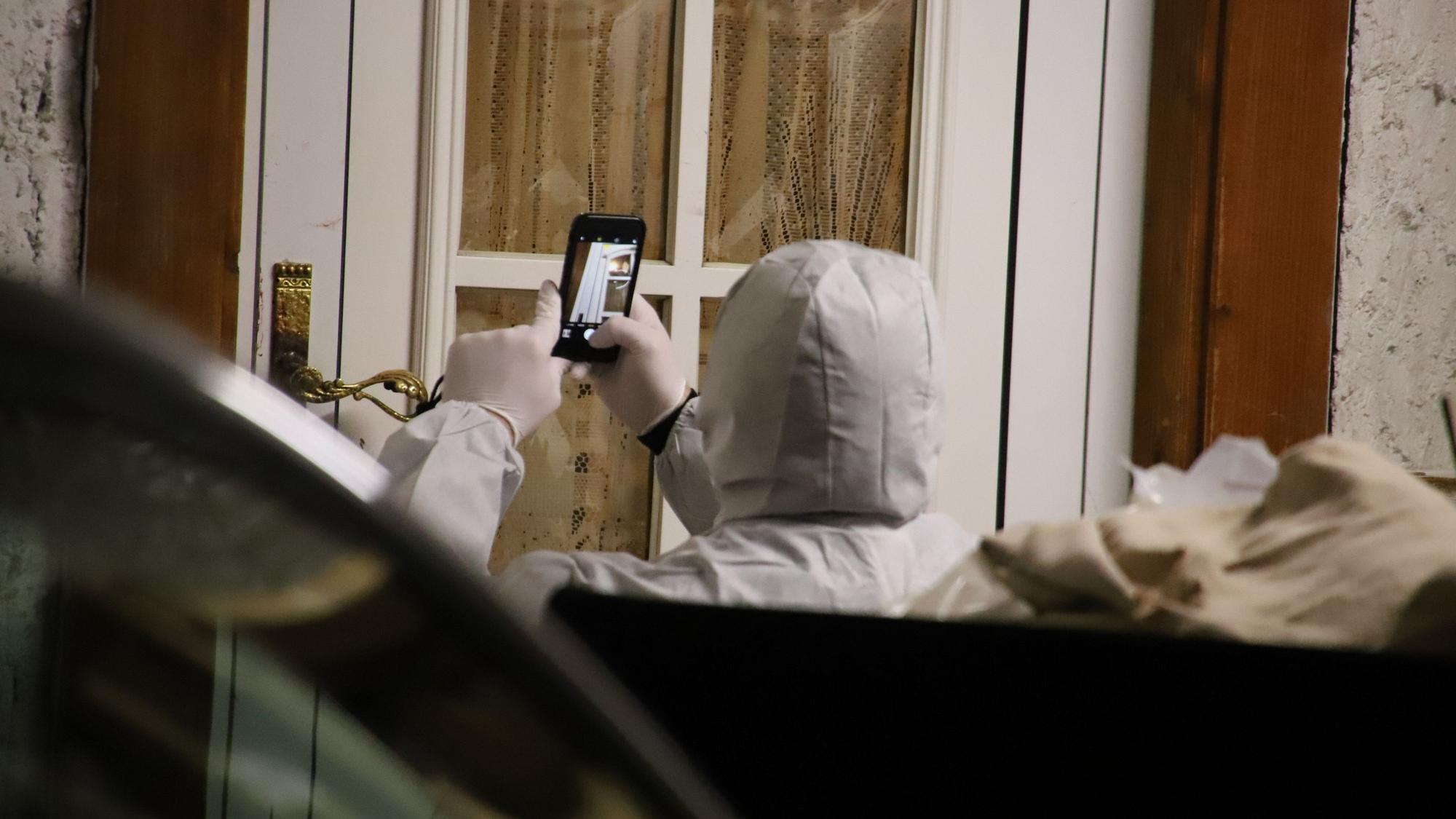 Eine Person in einer weißen Schutzkleidung macht Fotos von einer Eingangstür mit dem Handy.