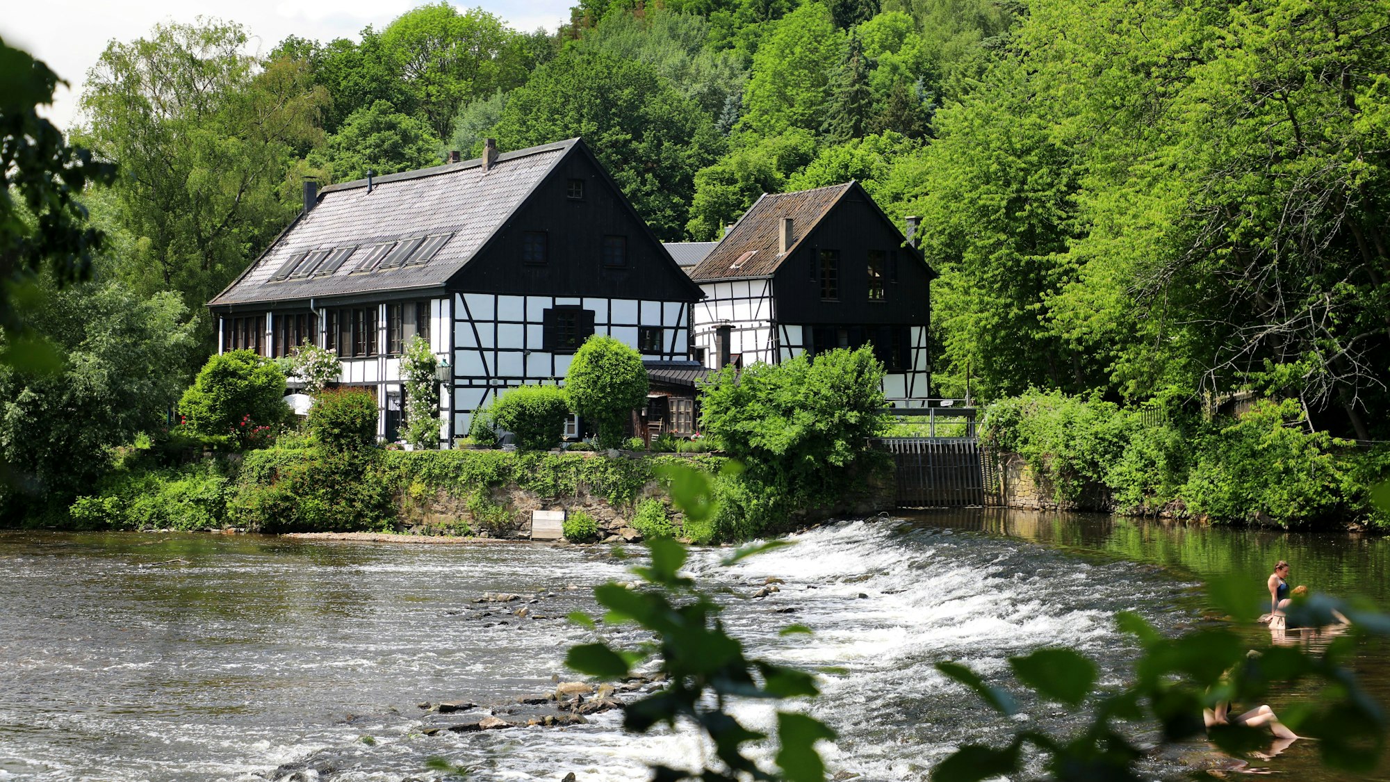 Ein Schleifkotten in Fachwerkbauweise steht an der Stromschnelle eines Stauwehrs in der Wupper.