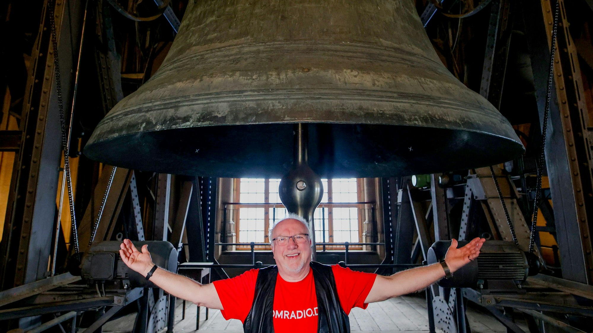Karnevalist und Priester Willibert Pauels steht unter der Petersglocke im Dom und breitet die Arme aus.