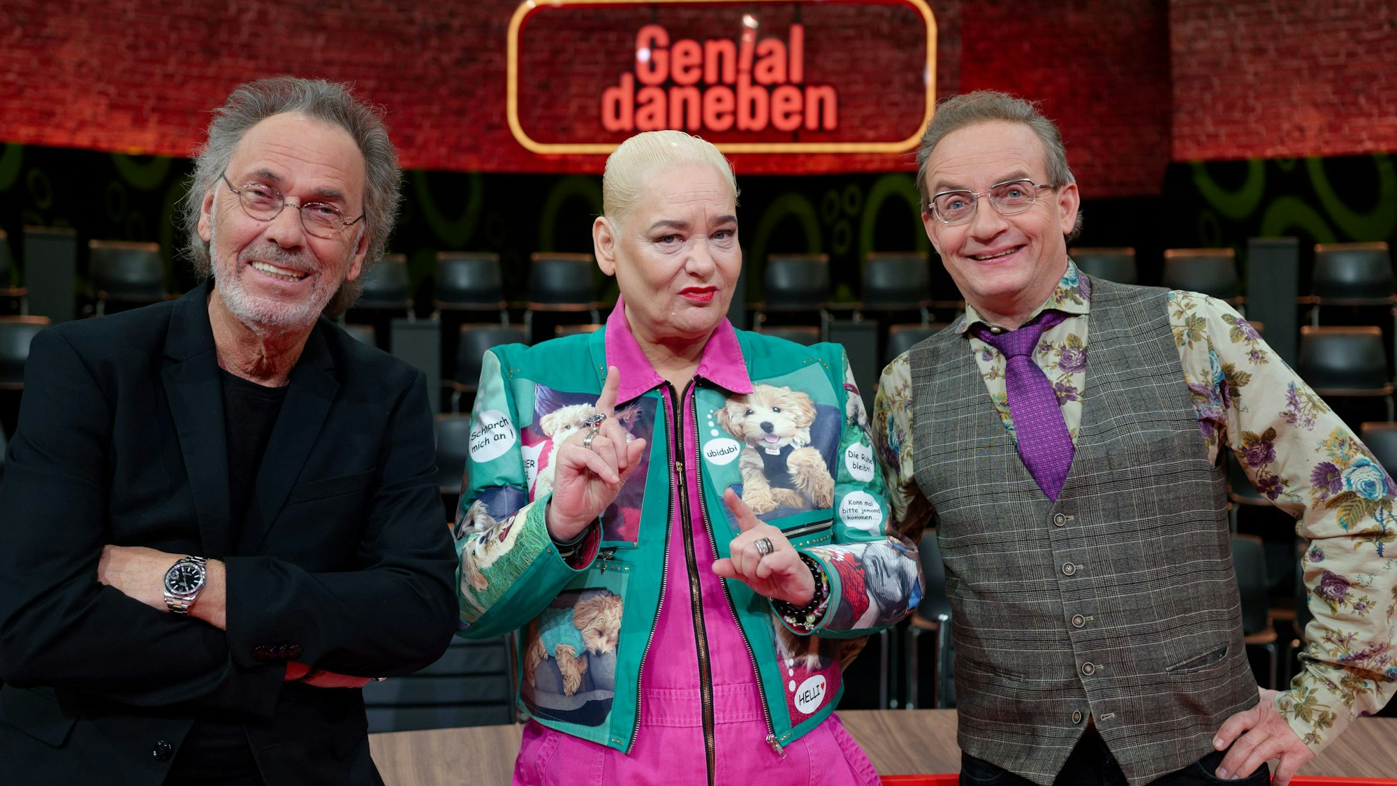 Hella von Sinnen steht am Set der Kultshow „Genial Daneben“ zwischen Hugo Egon Balder (l.) und Wigald Boning (r.). Alle drei posieren für die Kamera.