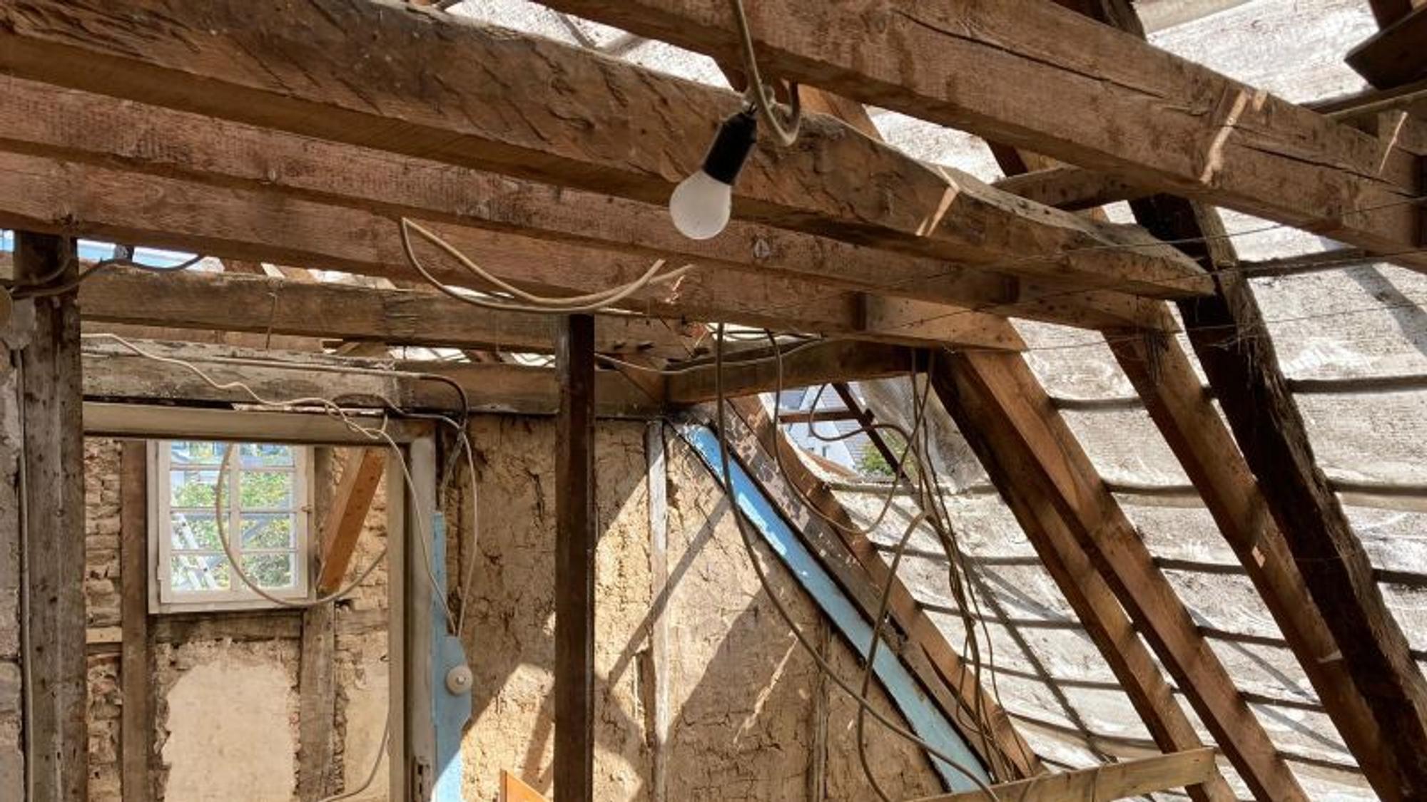 Es ist das noch nicht sanierte Dachgeschoss zu sehen. Von einem Holzbalken baumelt an einem Seil eine Glühbirne herunter.