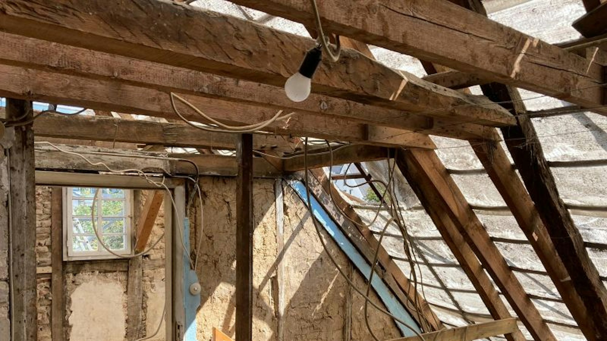 Es ist das noch nicht sanierte Dachgeschoss zu sehen. Von einem Holzbalken baumelt an einem Seil eine Glühbirne herunter.