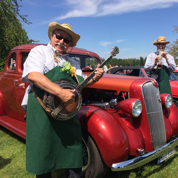 Es sind drei Männer zu sehen, die vor einem Oldtimer Musik spielen.
