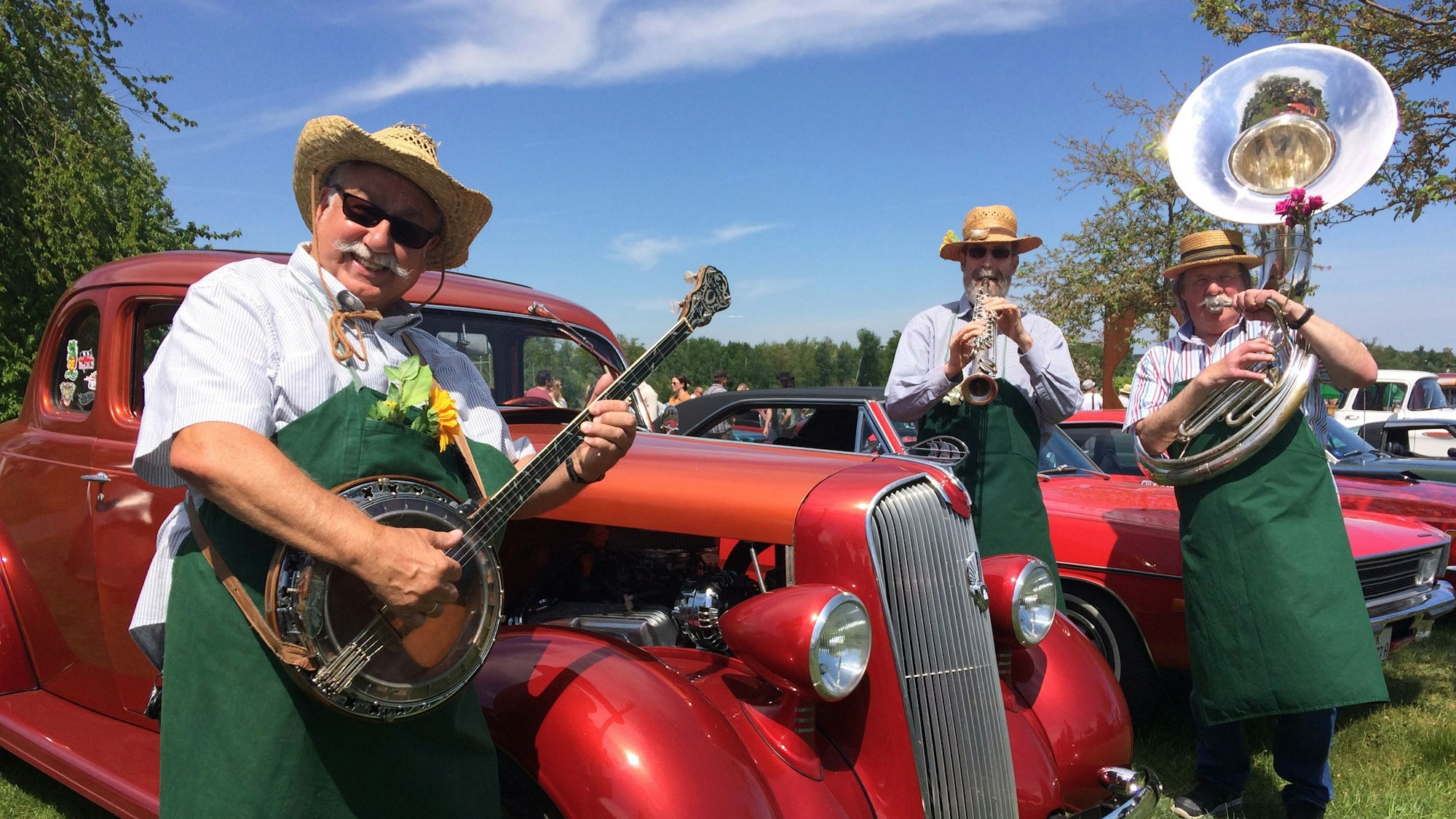 Es sind drei Männer zu sehen, die vor einem Oldtimer Musik spielen.