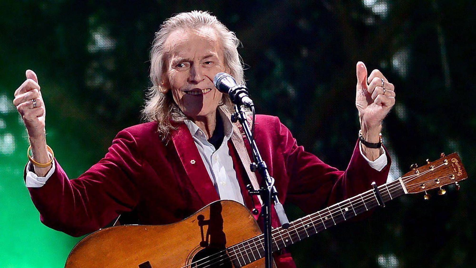 Gordon Lightfoot mit Gitarre auf der Bühne. Er hebt beide Daumen hoch und lacht.