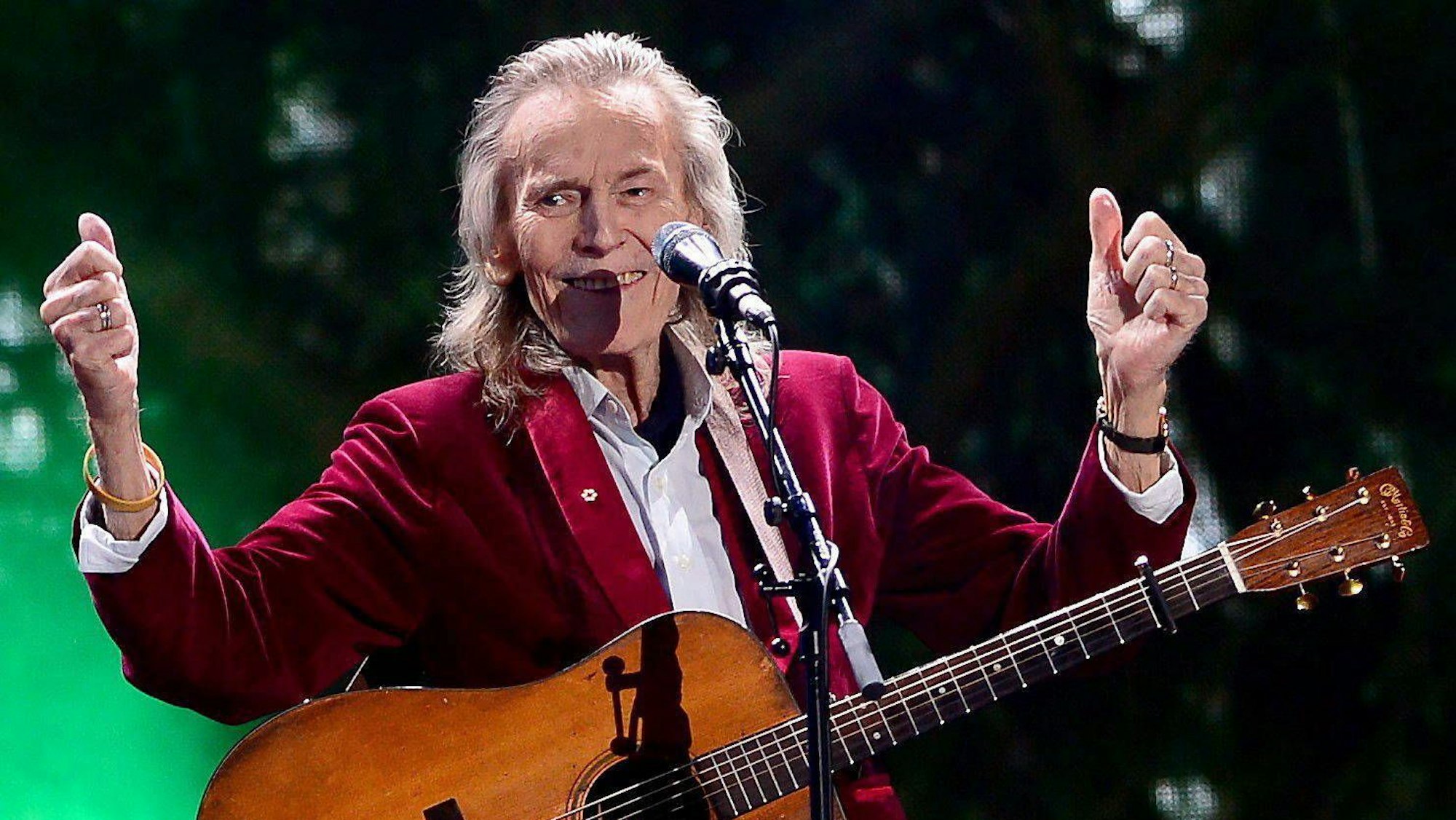 Gordon Lightfoot mit Gitarre auf der Bühne. Er hebt beide Daumen hoch und lacht.
