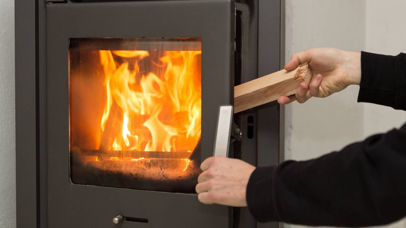 Ein Mann legt in seiner Wohnung ein Stück Holz in einen Kaminofen, in dem ein Feuer brennt.