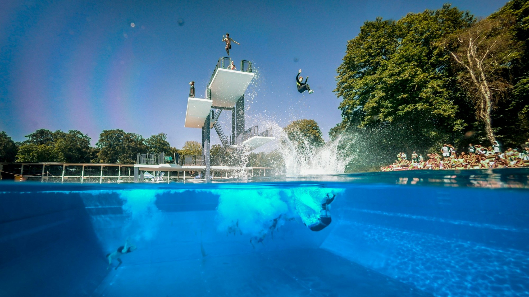 Menschen springen vom Sprungturm im Freibad Stadionbad in Köln ins Wasser.