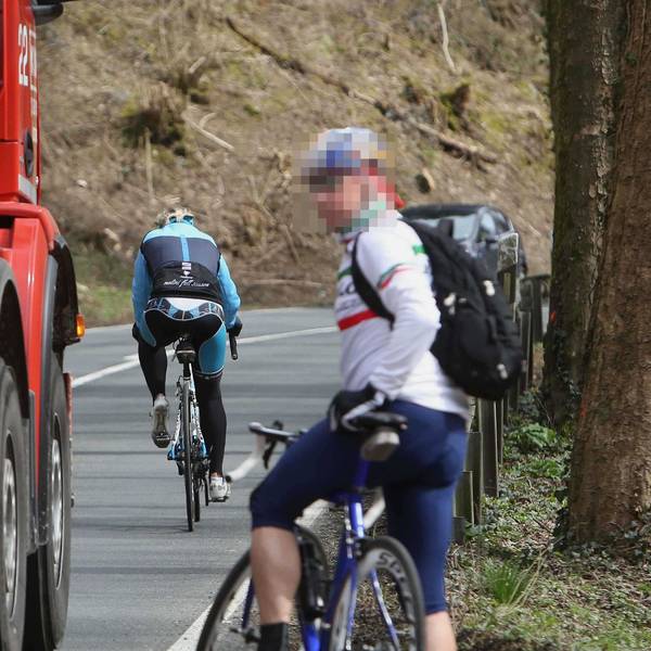 Ein Lastwagen fährt nahe an einem an Straßenrand stehenden Rennradfahrer vorbei; vor dem Lkw fährt ein weiterer Radfahrer.