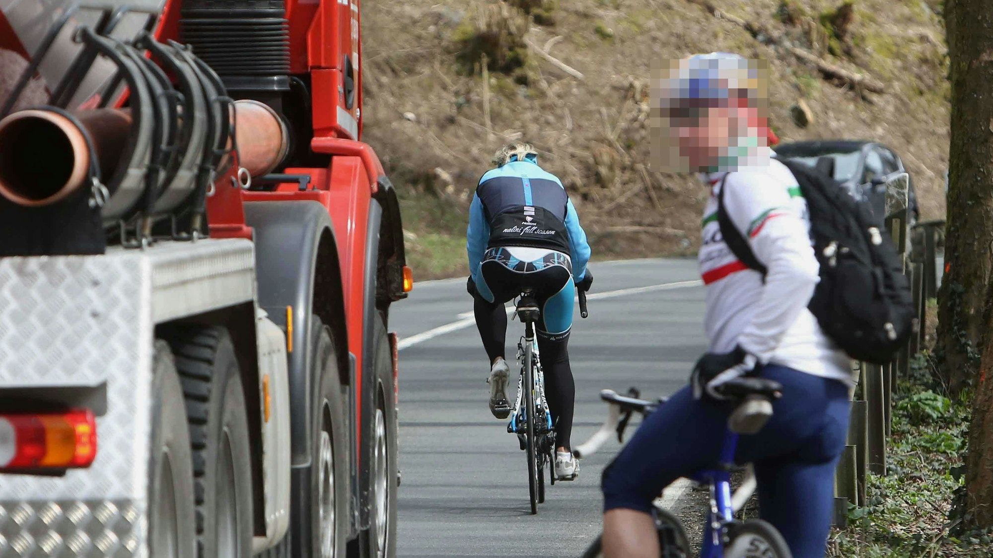 Ein Lastwagen fährt nahe an einem an Straßenrand stehenden Rennradfahrer vorbei; vor dem Lkw fährt ein weiterer Radfahrer.