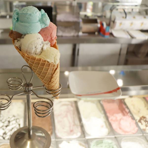 Eine große Eiswaffel mit mehreren Eiskugeln ist mittig im Bild platziert. Sie befindet sich auf einem Eiswaffelständer an der Theke eines Eiscafés.
