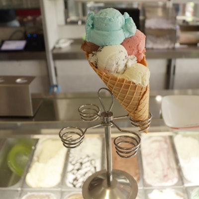 Eine große Eiswaffel mit mehreren Eiskugeln ist mittig im Bild platziert. Sie befindet sich auf einem Eiswaffelständer an der Theke eines Eiscafés.