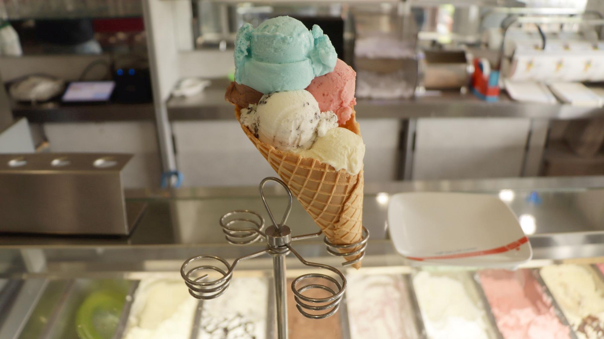 Eine große Eiswaffel mit mehreren Eiskugeln ist mittig im Bild platziert. Sie befindet sich auf einem Eiswaffelständer an der Theke eines Eiscafés.