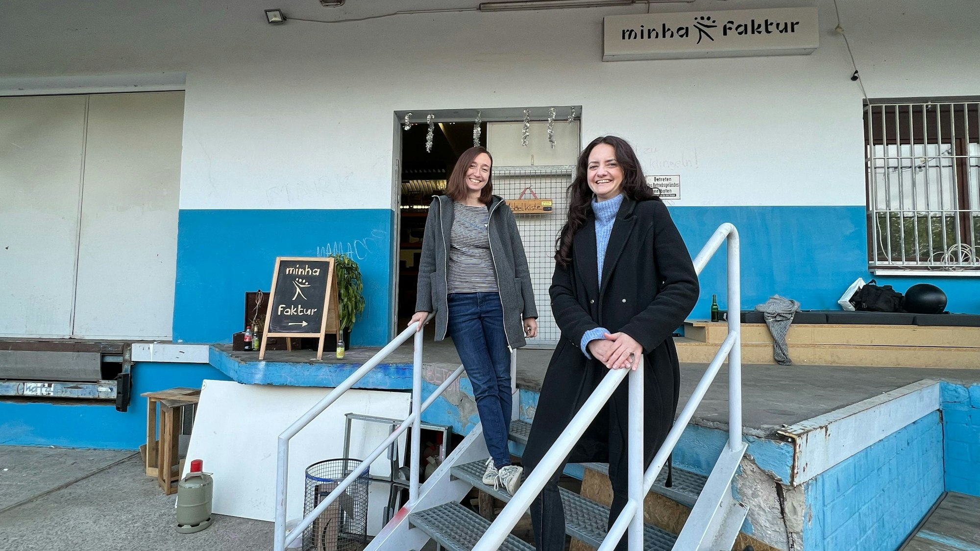 Zwei junge Frauen stehen auf einer Treppe, die zu einer Lagerhalle führt.
