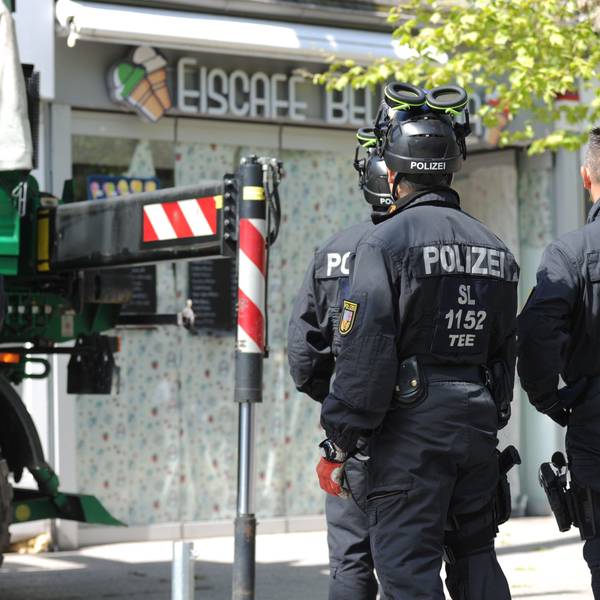 Polizisten sichern im Rahmen einer Razzia Beweismittel. Zu sehen sind Einsatzkräfte, auf deren Rücken "Polizei" zu lesen ist. Sei stehen vor einem Eiscafé.