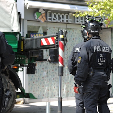 Polizisten sichern im Rahmen einer Razzia Beweismittel. Zu sehen sind Einsatzkräfte, auf deren Rücken "Polizei" zu lesen ist. Sei stehen vor einem Eiscafé.