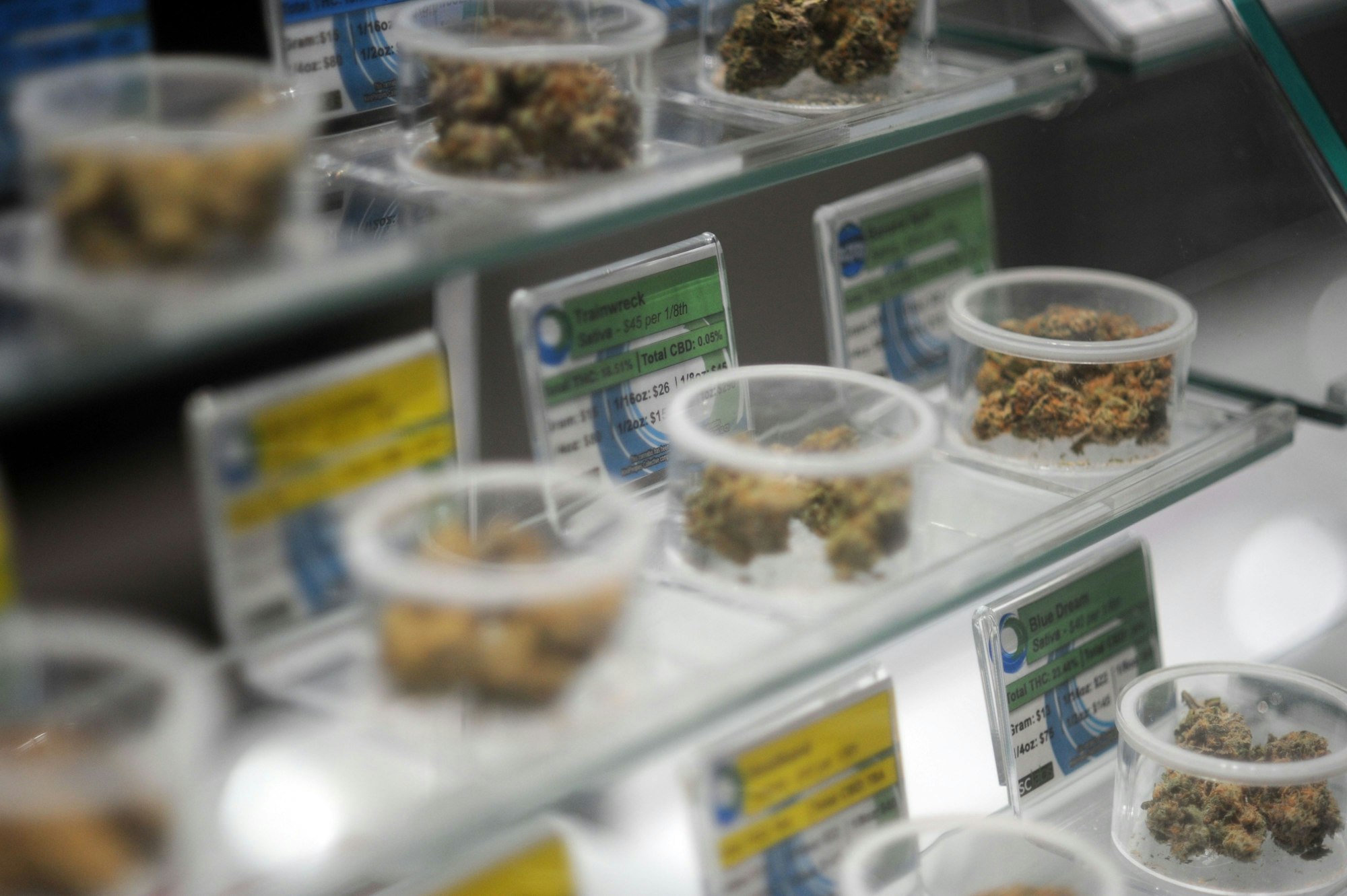 Auf Glasregalen stehen durchsichtige Plastikschalen, die mit Marihuana-Blüten gefüllt sind. Hinter den Schalen geben gelbe und grüne Schilder Auskunft über Eigenschaften und Preis der Waren.