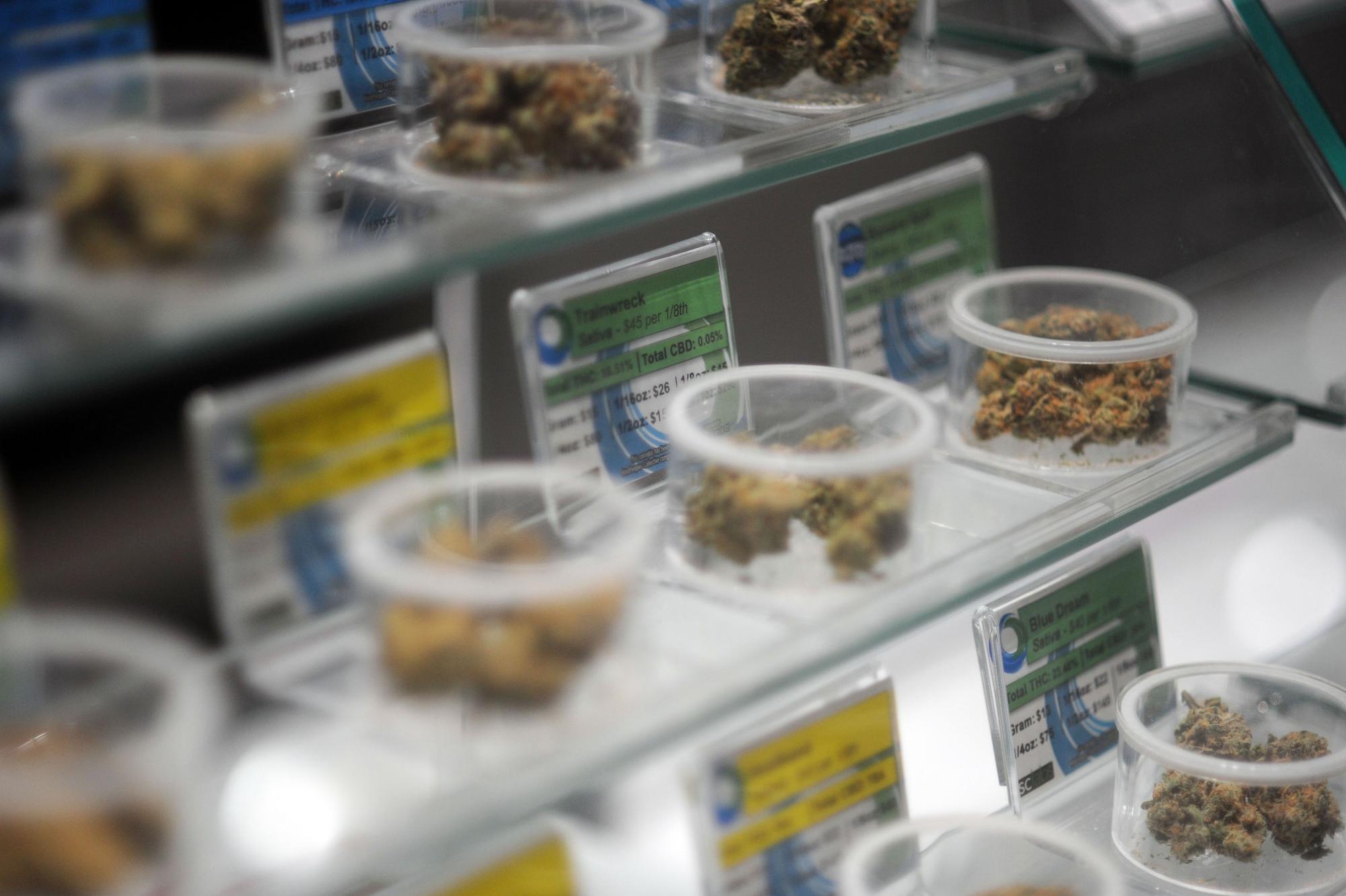 Auf Glasregalen stehen durchsichtige Plastikschalen, die mit Marihuana-Blüten gefüllt sind. Hinter den Schalen geben gelbe und grüne Schilder Auskunft über Eigenschaften und Preis der Waren.