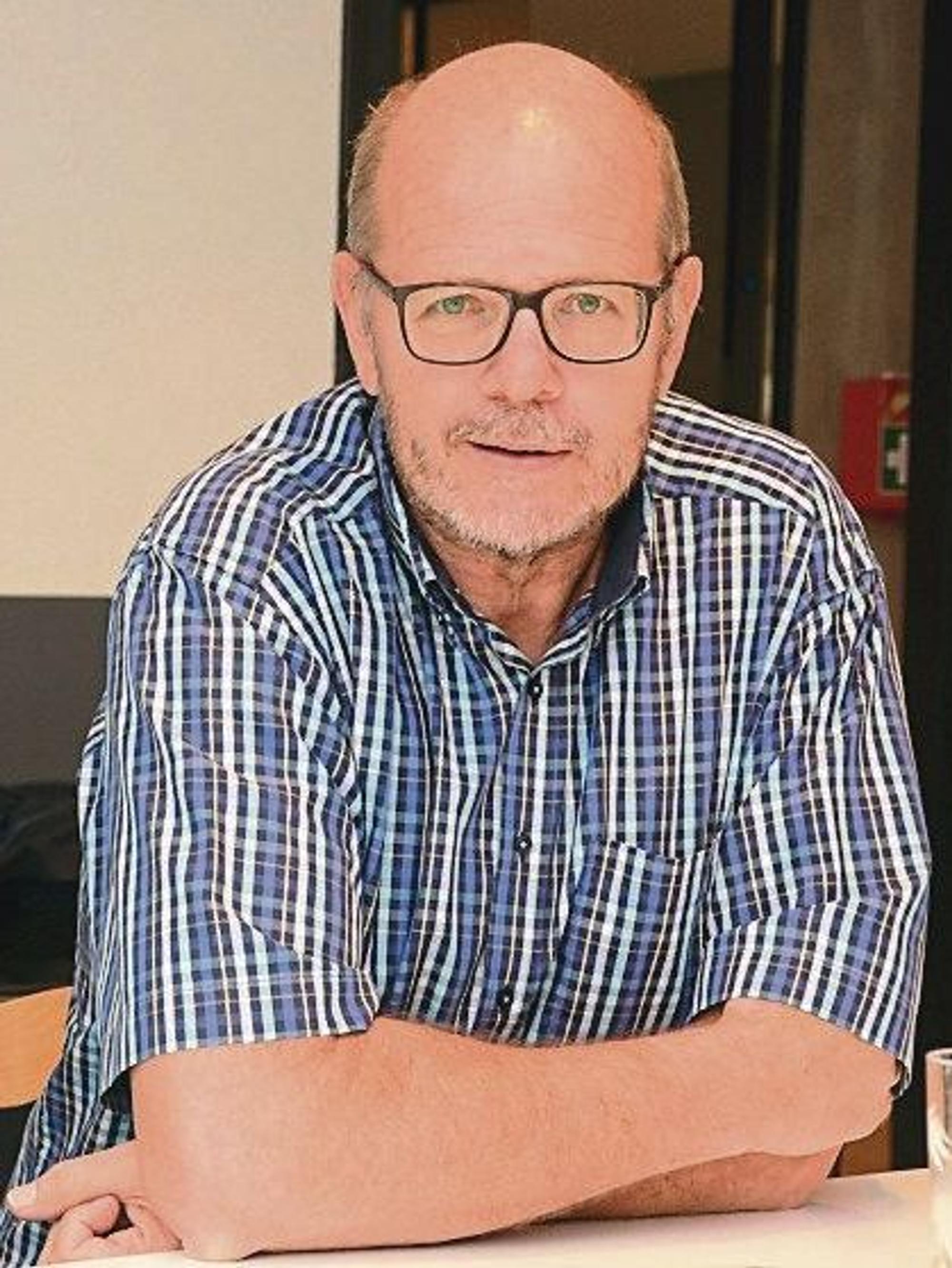 Ein Mann mit Glatze und Brille; er trägt ein kariertes Kurzarmhemd.