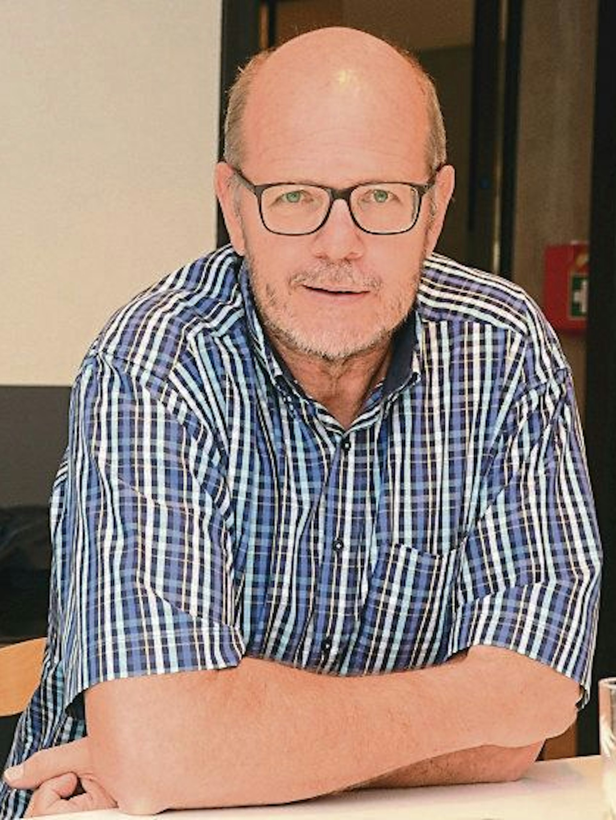 Ein Mann mit Glatze und Brille; er trägt ein kariertes Kurzarmhemd.