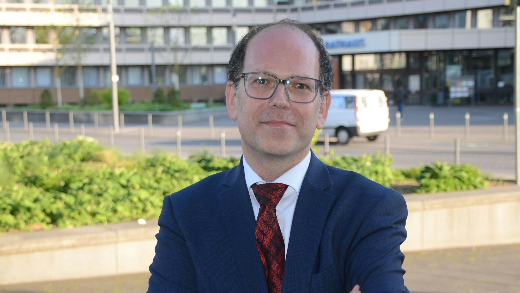 Mit verschränkten Armen steht ein Mann vor dem Troisdorfer Rathaus. Er trägt eine Brille, einen blauen Anzug und eine rote Krawatte.
