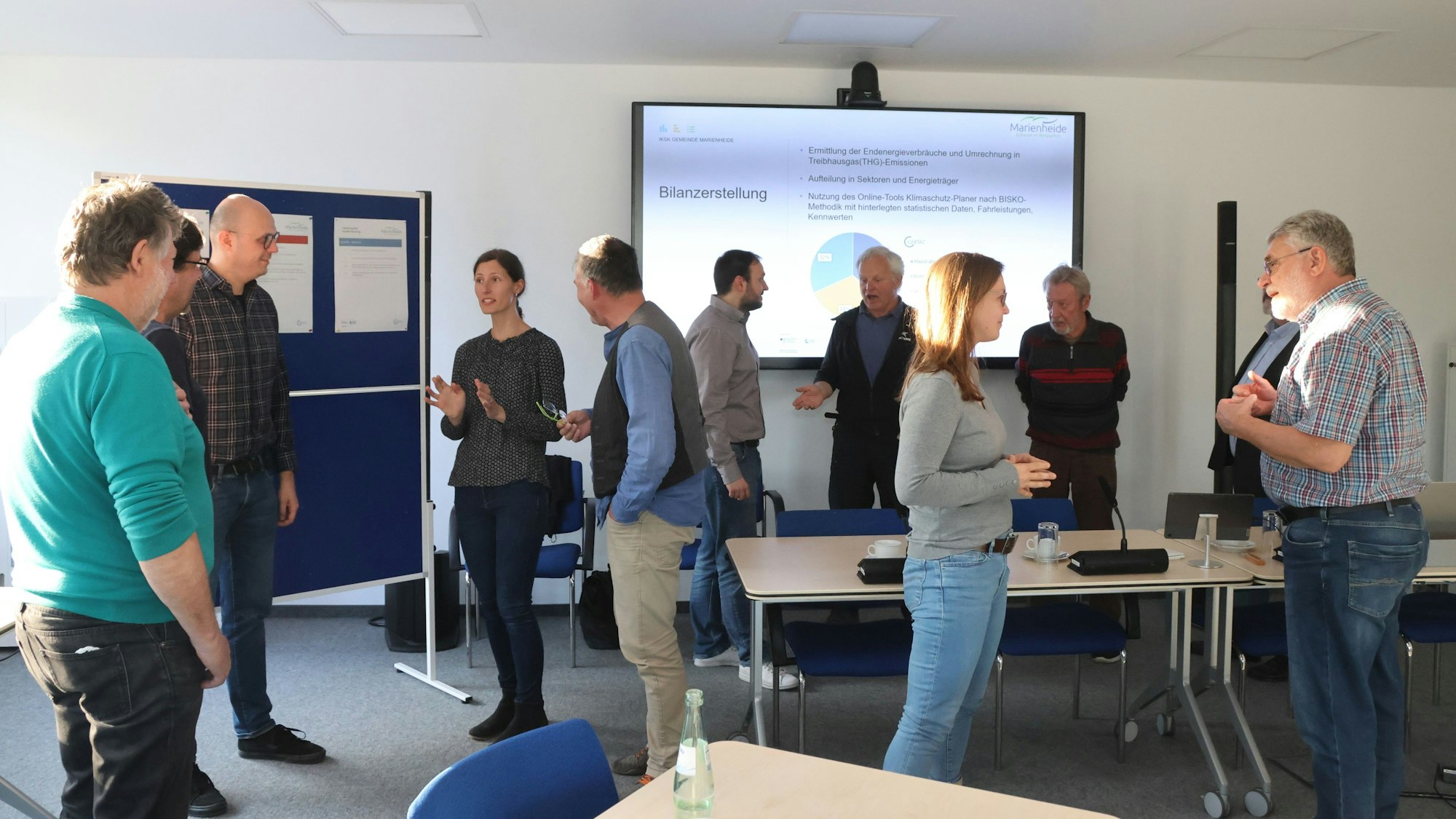 Mehrere Männer und Frauen stehen in einem Raum verteilt und diskutieren in Gruppen miteinander. Im Hintergrund ist auf einer Leinwand eine Präsentation zum Klimaschutzkonzept zu sehen.
