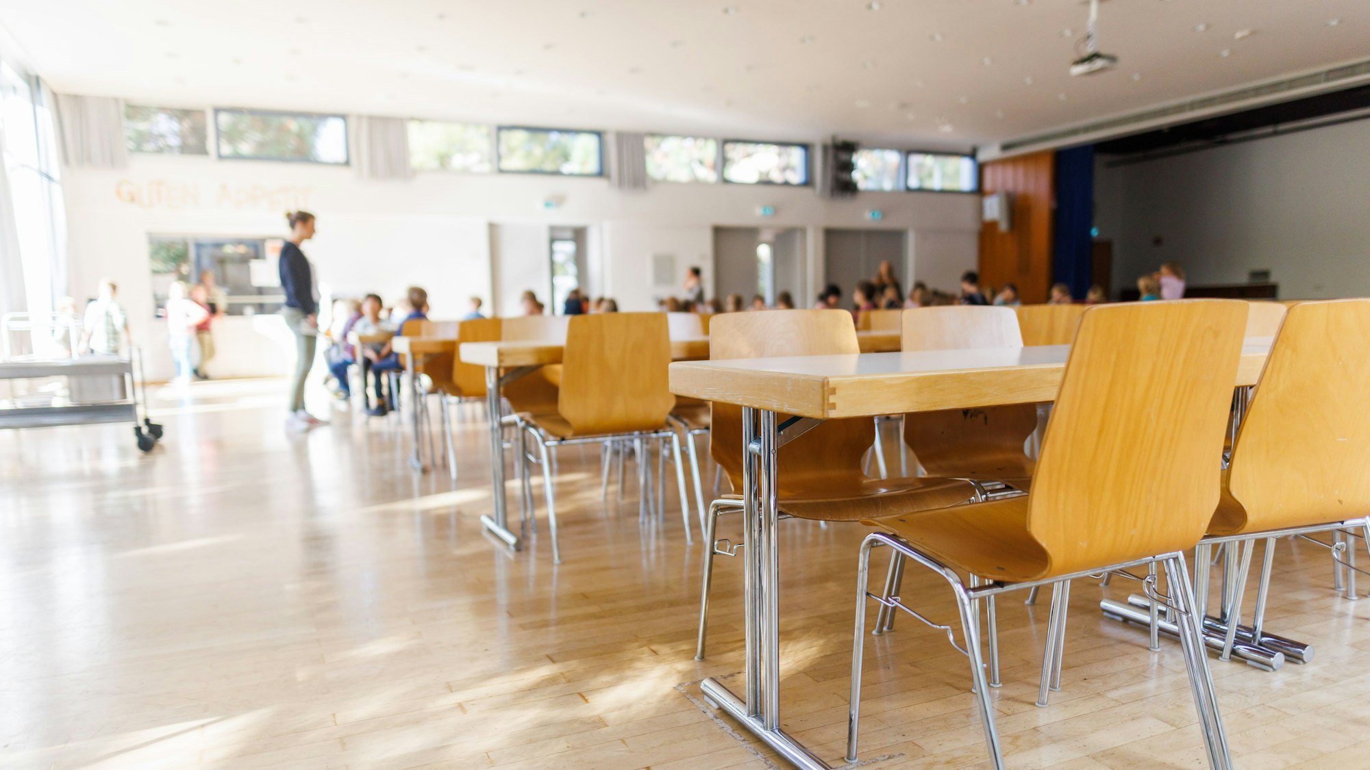 Stühle und Tische für Erwachsene stehen in einer zur Schulmensa umfunktionierten Aula einer Grundschule während im Hintergrund eine Lehrerin und Schüler zu sehen sind.