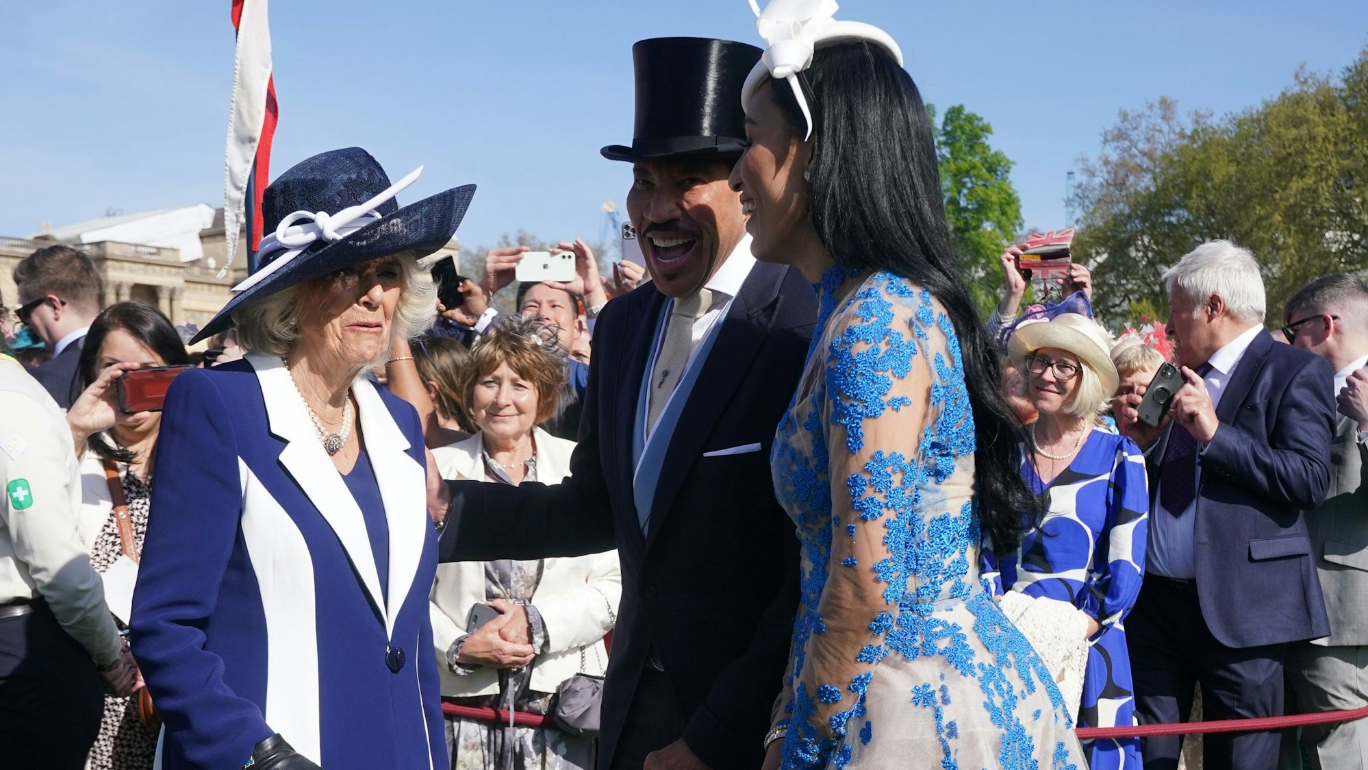 Königin Camilla, Lionel Richie und Lisa Parigi unterhalten sich im Garten, dahinter Schaulustige.