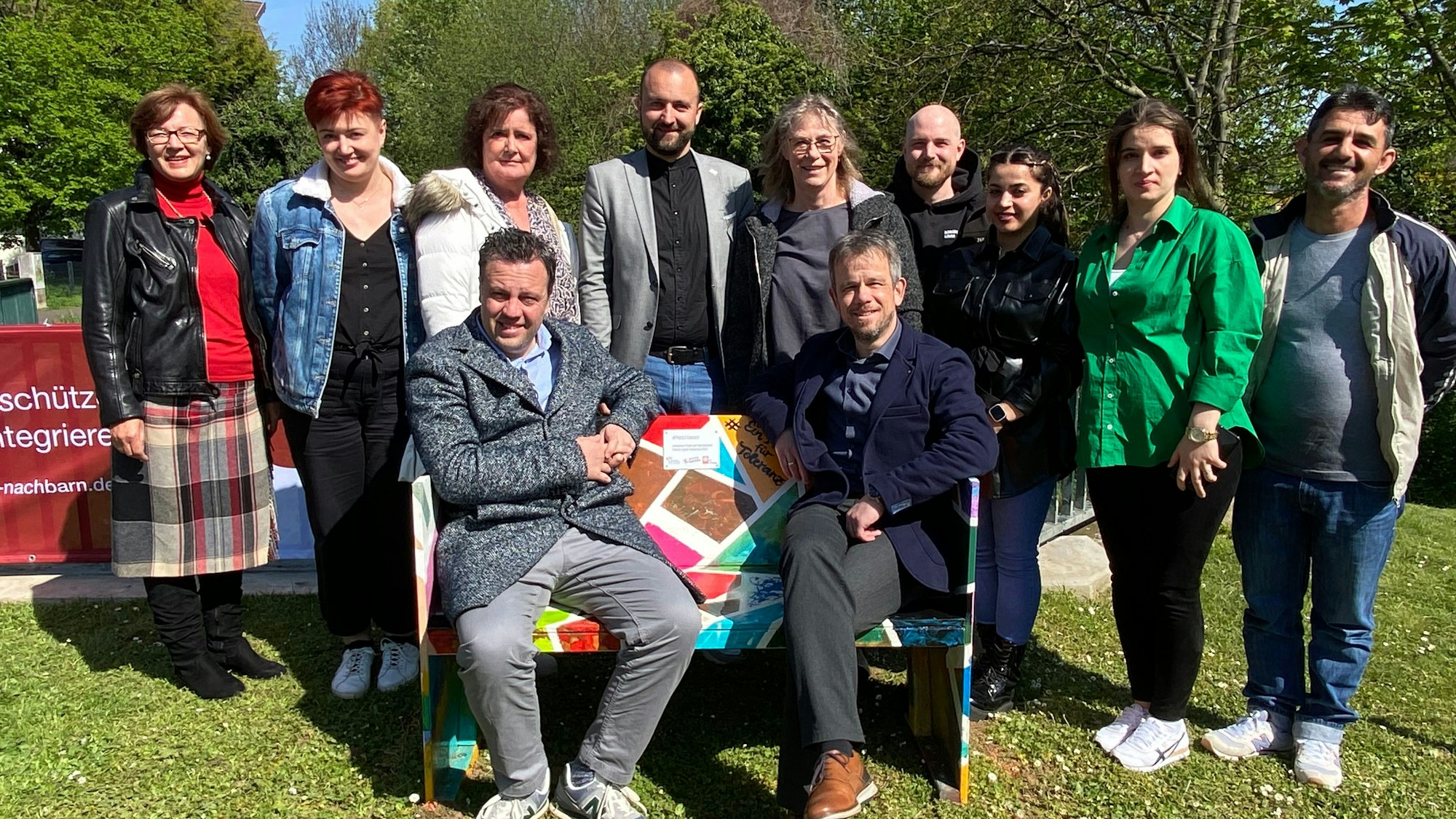 Es sind mehrere Menschen zu sehen, die um die Bank herumstehen. Zwei von ihnen sitzen auf ihr drauf.