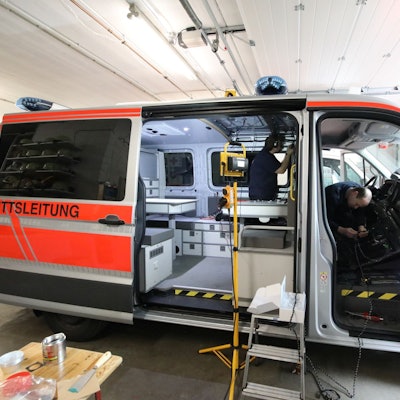 Ein Transporter steht mit offener Schiebetür und geöffneter Beifahrertür in einer Halle. Zwei Männer arbeiten in dem Fahrzeug, das durch einen Scheinwerfer ausgeleuchtet wird.