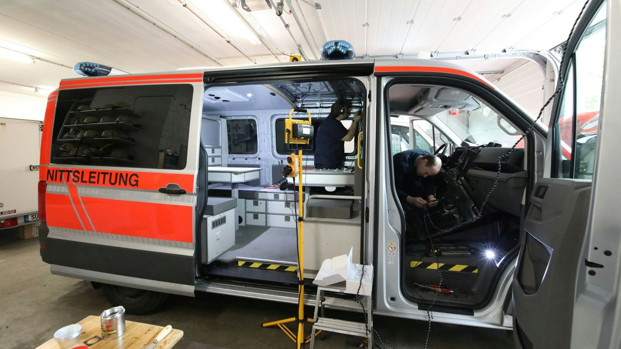 Ein Transporter steht mit offener Schiebetür und geöffneter Beifahrertür in einer Halle. Zwei Männer arbeiten in dem Fahrzeug, das durch einen Scheinwerfer ausgeleuchtet wird.