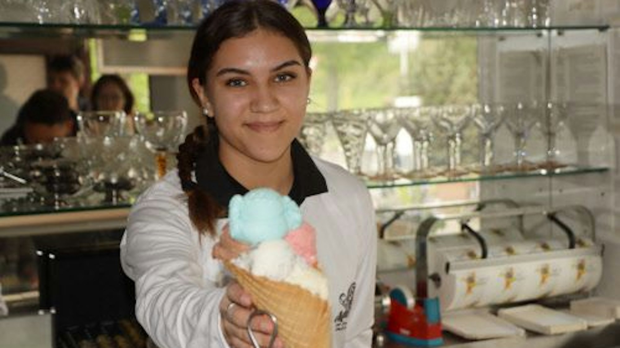 Eine Mitarbeiterin der Eisdiele MammaMia in Wesseling-Berzdorf hält eine große Eiswaffel mit mehreren Kugeln Eis in Richtung der Kamera und lächelt dabei.