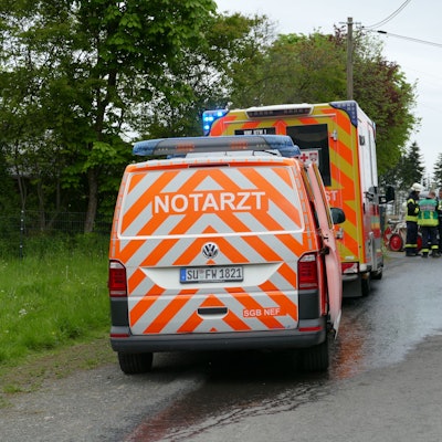 Ein Notarztwagen und ein Krankenwagen stehen auf einer asphaltierten Zufahrt. Im Hintergrund sind Feuerwehrleute und Rettungskräfte.